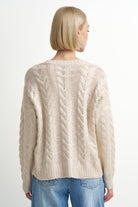 A woman with a blonde bob is shown from the back wearing a cream-colored cable-knit sweater and blue jeans against a plain white background.