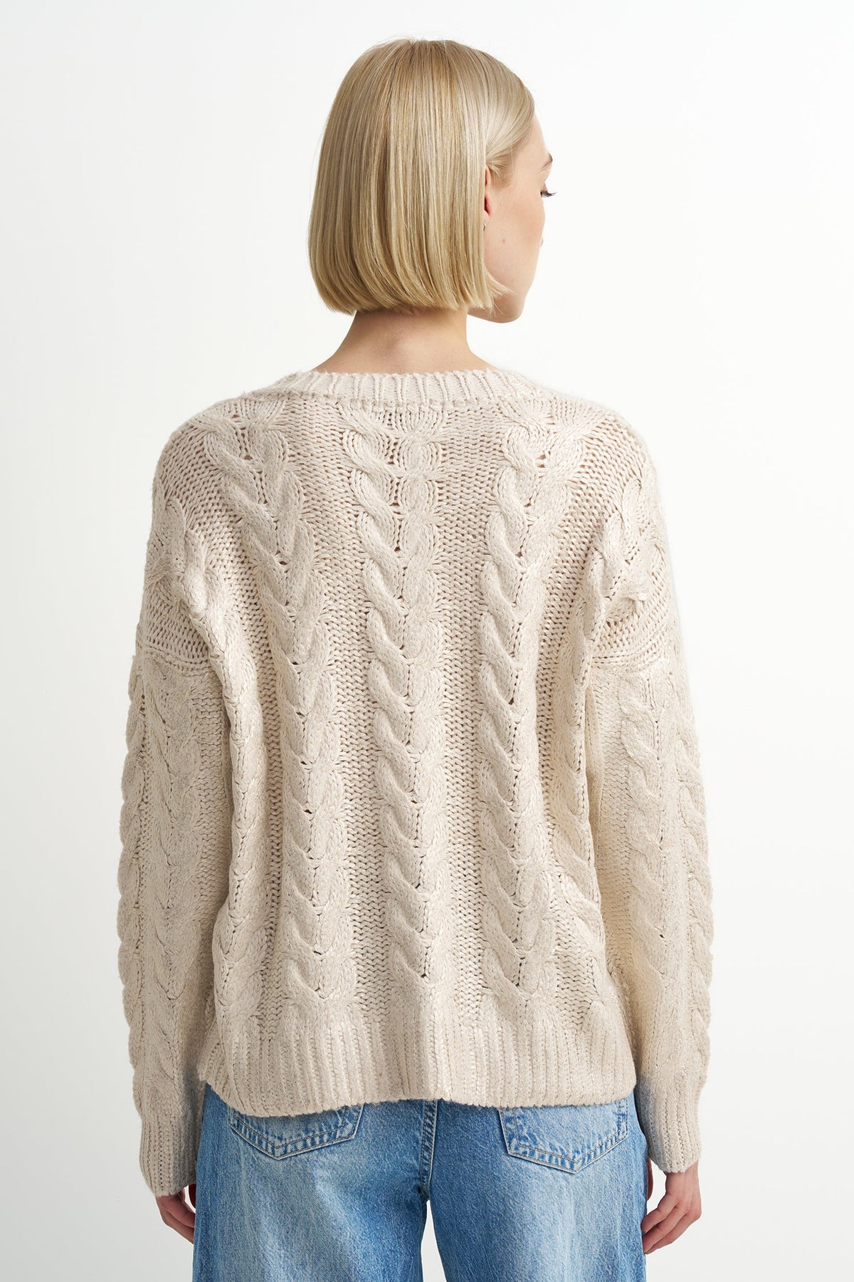 A woman with a blonde bob is shown from the back wearing a cream-colored cable-knit sweater and blue jeans against a plain white background.
