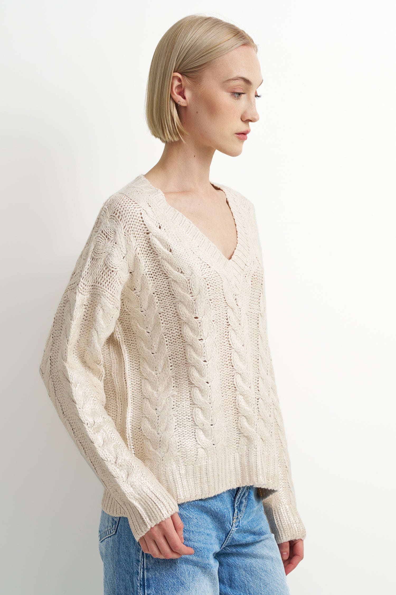 A woman with short blonde hair wearing a cream-colored cable-knit V-neck sweater and blue jeans, shown in a side profile against a plain white studio background.