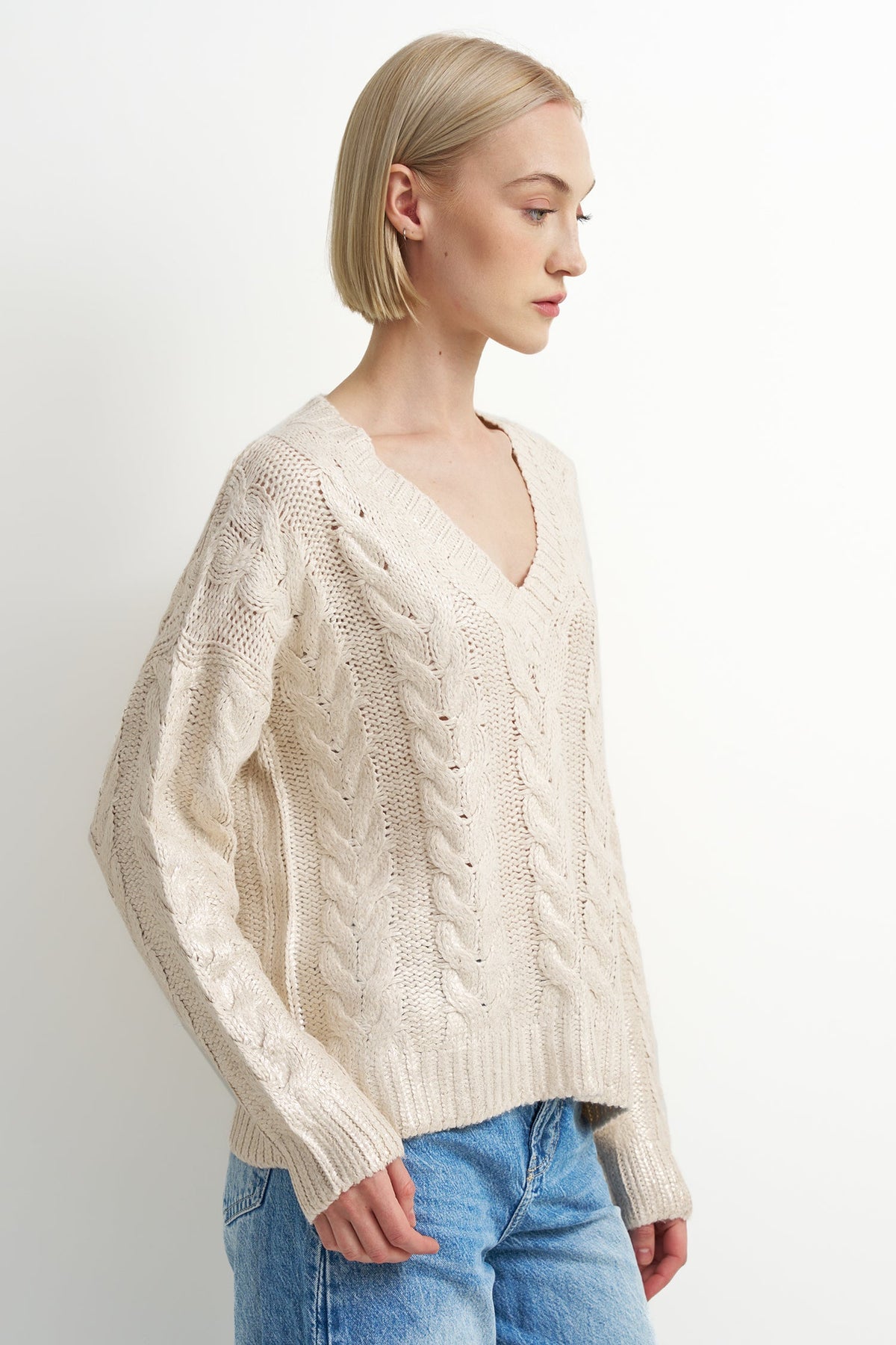 A woman with short blonde hair wearing a cream-colored cable-knit V-neck sweater and blue jeans, shown in a side profile against a plain white studio background.