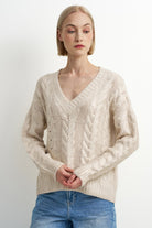A woman wearing a cream-colored cable-knit V-neck sweater and blue jeans, standing against a plain light gray studio background.