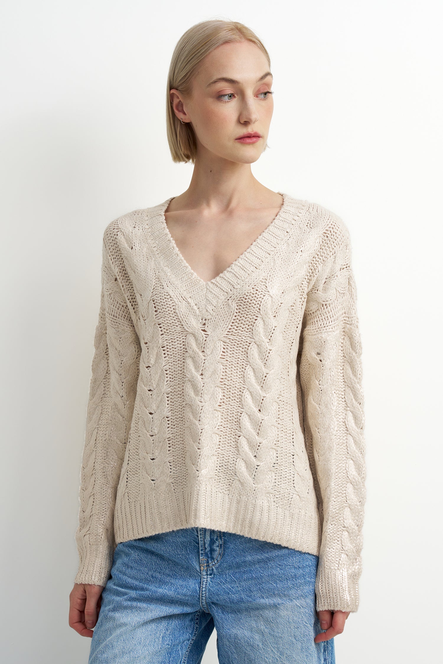 A woman wearing a cream-colored cable-knit V-neck sweater and blue jeans, standing against a plain light gray studio background.